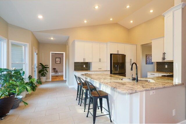 kitchen with an island with sink, stainless steel fridge, light stone countertops, white cabinets, and tasteful backsplash