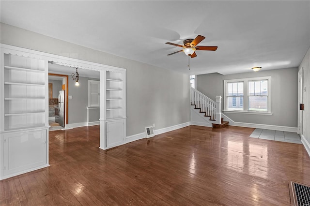 unfurnished living room with built in shelves, ceiling fan with notable chandelier, and dark hardwood / wood-style flooring