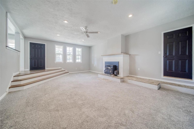 unfurnished living room with carpet flooring, ceiling fan, a wood stove, and a textured ceiling