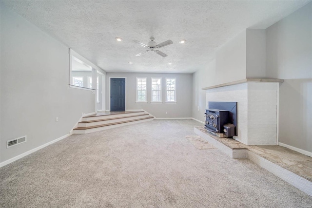 unfurnished living room with a wood stove, ceiling fan, carpet floors, and a textured ceiling