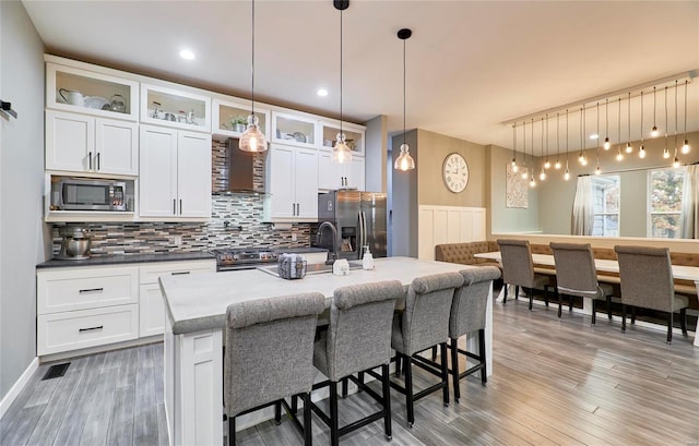 kitchen featuring stainless steel appliances, wall chimney exhaust hood, an island with sink, backsplash, and pendant lighting