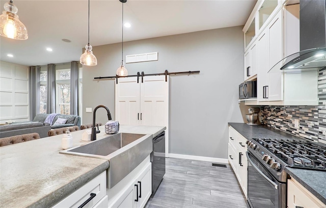 kitchen with white cabinets, wall chimney exhaust hood, black appliances, a barn door, and decorative light fixtures