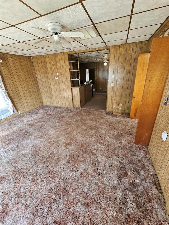 unfurnished room featuring dark colored carpet, ceiling fan, a drop ceiling, and wood walls