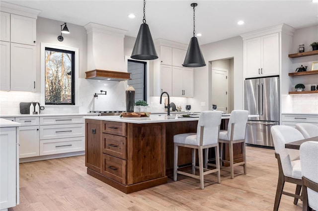 kitchen featuring a center island with sink, white cabinetry, light hardwood / wood-style floors, high end fridge, and custom range hood