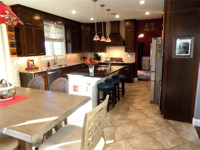 kitchen featuring stainless steel appliances, light stone counters, custom range hood, a kitchen island with sink, and pendant lighting