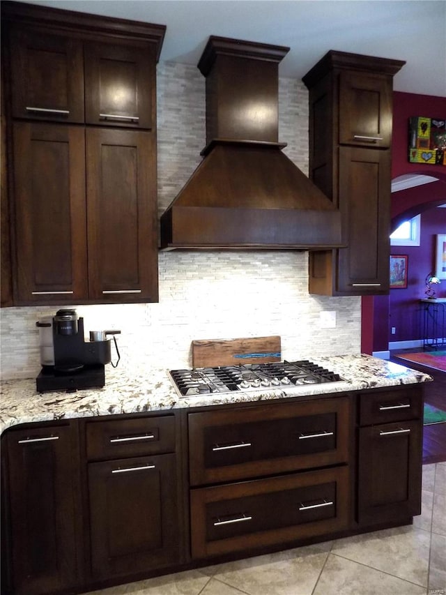 kitchen with light stone counters, light tile patterned flooring, custom range hood, dark brown cabinets, and decorative backsplash