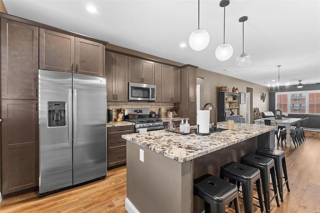 kitchen featuring an island with sink, light hardwood / wood-style flooring, stainless steel appliances, light stone countertops, and decorative light fixtures