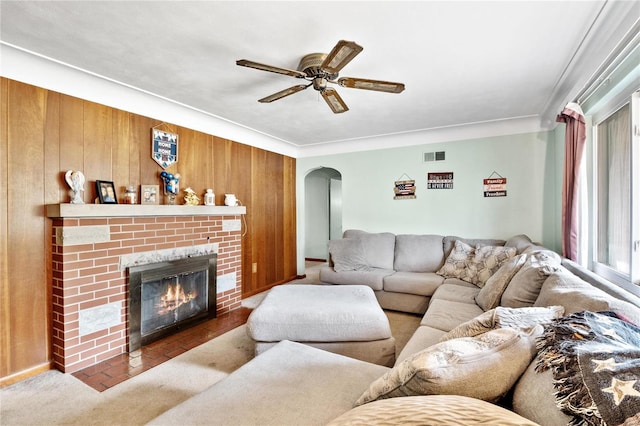 living room with a brick fireplace, ceiling fan, wooden walls, and crown molding