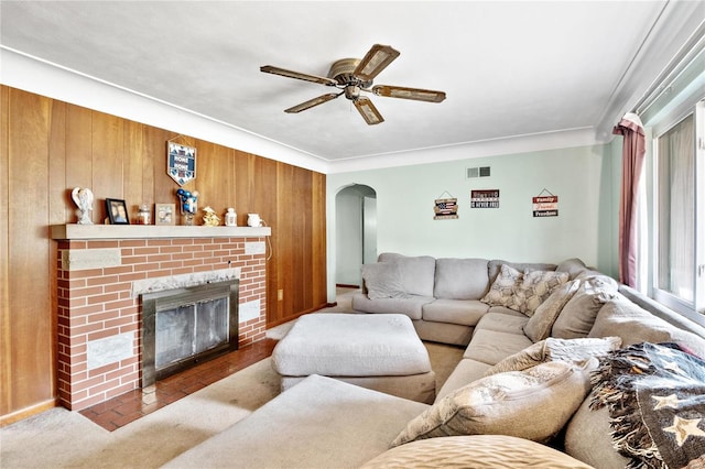 living room with a brick fireplace, ceiling fan, wooden walls, and ornamental molding