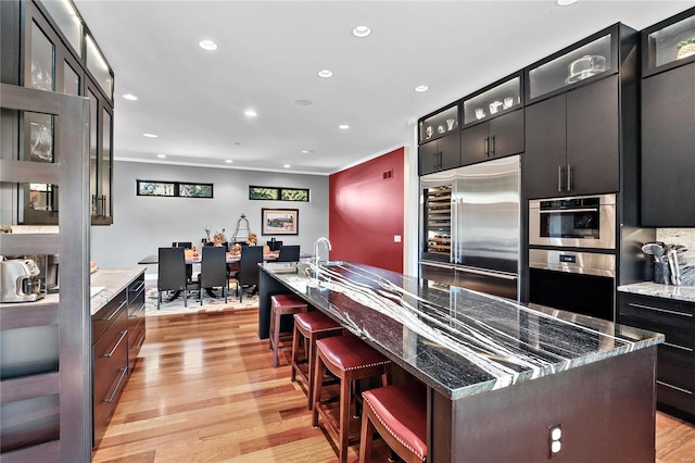 kitchen with stainless steel appliances, dark stone counters, a center island with sink, a kitchen breakfast bar, and light hardwood / wood-style flooring