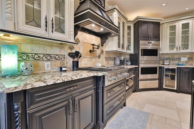 kitchen with appliances with stainless steel finishes, light stone counters, premium range hood, and beverage cooler