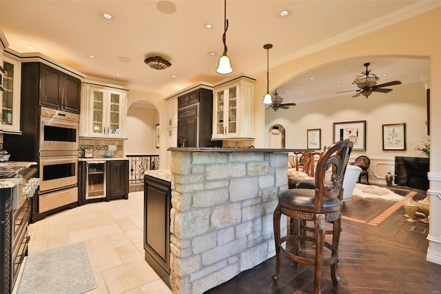 kitchen with hanging light fixtures, ceiling fan, ornamental molding, double oven, and beverage cooler