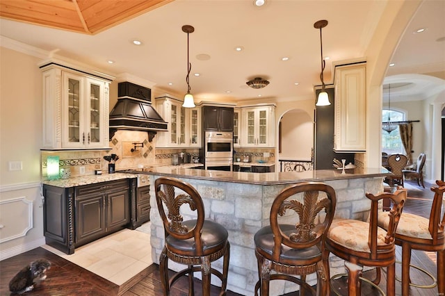 bar with stainless steel double oven, premium range hood, pendant lighting, dark stone counters, and light tile patterned flooring