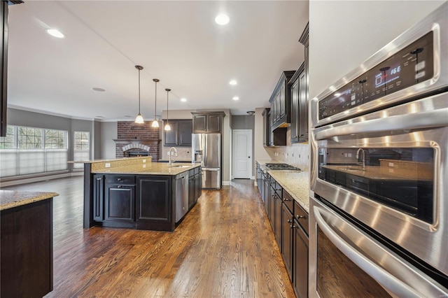 kitchen with dark wood-type flooring, hanging light fixtures, stainless steel appliances, tasteful backsplash, and an island with sink