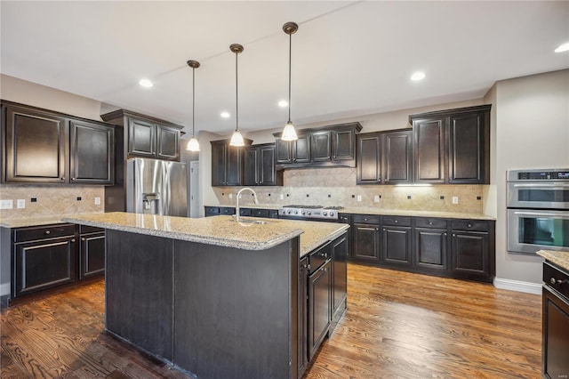 kitchen with stainless steel appliances, a kitchen island with sink, dark wood-type flooring, sink, and pendant lighting