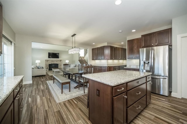 kitchen with a stone fireplace, dark brown cabinetry, dark wood-type flooring, stainless steel fridge, and pendant lighting