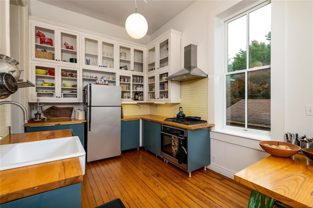 kitchen featuring stainless steel appliances, white cabinetry, backsplash, hanging light fixtures, and wall chimney range hood