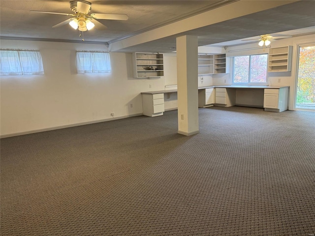 basement featuring built in shelves, dark carpet, ornamental molding, ceiling fan, and built in desk