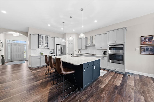 kitchen with sink, decorative light fixtures, an island with sink, stainless steel appliances, and wall chimney range hood