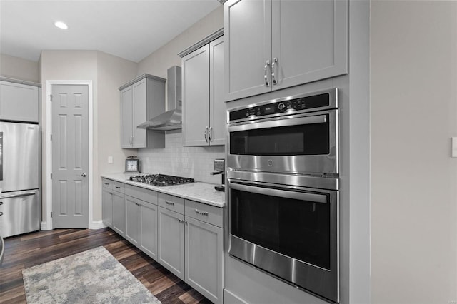 kitchen featuring wall chimney range hood, dark wood-type flooring, gray cabinetry, stainless steel appliances, and light stone counters