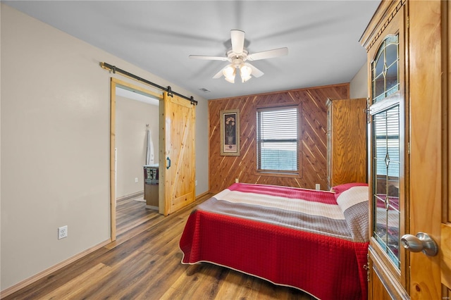 bedroom featuring a barn door, ceiling fan, wooden walls, and dark wood-type flooring