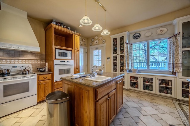 kitchen with sink, pendant lighting, white appliances, a kitchen island with sink, and custom exhaust hood