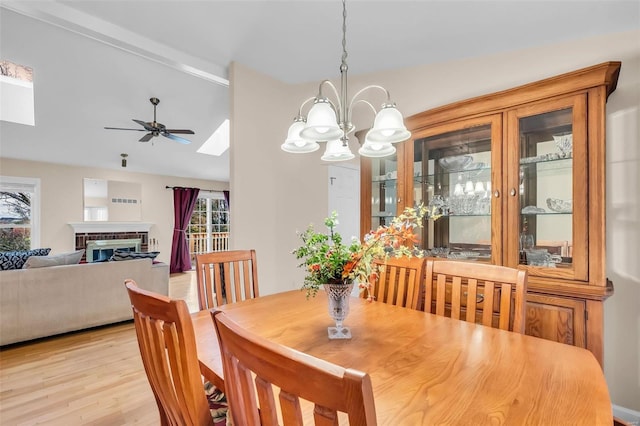 dining room featuring ceiling fan with notable chandelier, light wood-type flooring, a brick fireplace, and vaulted ceiling with skylight