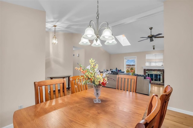 dining area featuring a brick fireplace, ceiling fan with notable chandelier, light hardwood / wood-style flooring, and vaulted ceiling with skylight