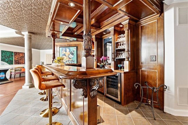 bar featuring coffered ceiling, wine cooler, decorative columns, light tile patterned floors, and ornamental molding