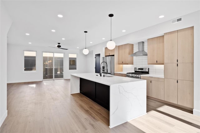 kitchen with wall chimney exhaust hood, hanging light fixtures, light hardwood / wood-style flooring, a center island with sink, and appliances with stainless steel finishes