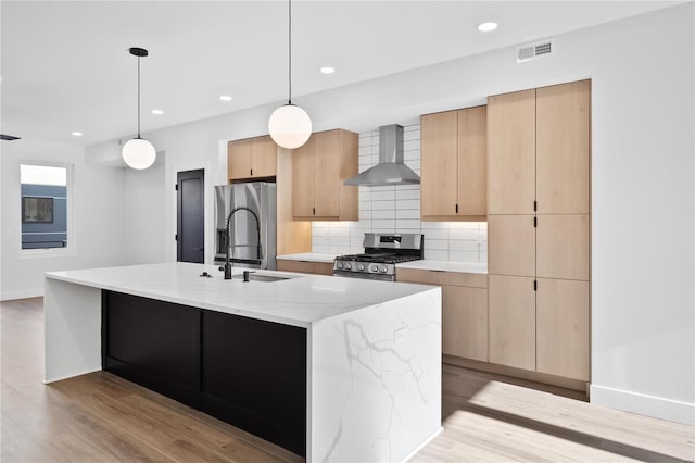 kitchen featuring hanging light fixtures, wall chimney exhaust hood, light wood-type flooring, an island with sink, and appliances with stainless steel finishes