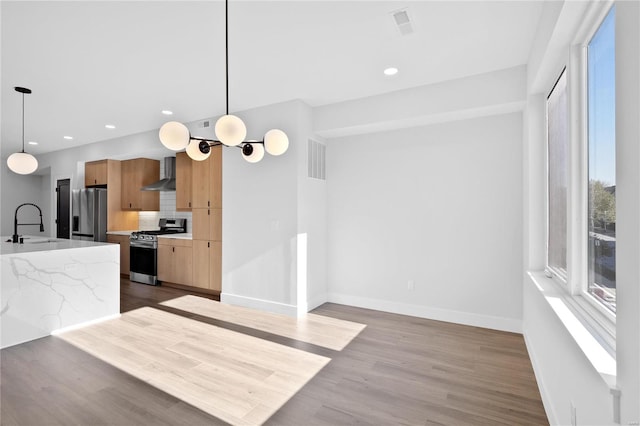 kitchen with wall chimney exhaust hood, hanging light fixtures, light stone counters, appliances with stainless steel finishes, and light wood-type flooring