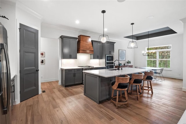 kitchen with gray cabinetry, premium range hood, an island with sink, light hardwood / wood-style floors, and decorative light fixtures