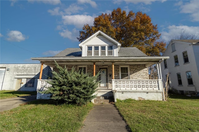 bungalow featuring a front yard, covered porch, stone siding, and a shingled roof