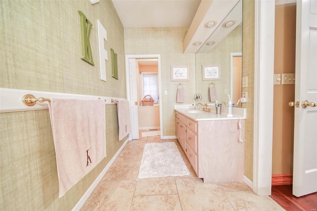 full bathroom featuring double vanity, wallpapered walls, baseboards, and a sink