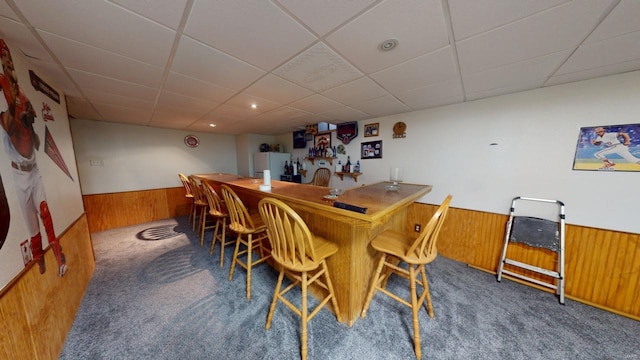 carpeted dining area featuring bar, a drop ceiling, and wooden walls