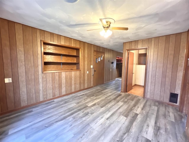 empty room with built in shelves, light hardwood / wood-style floors, ceiling fan, and wood walls