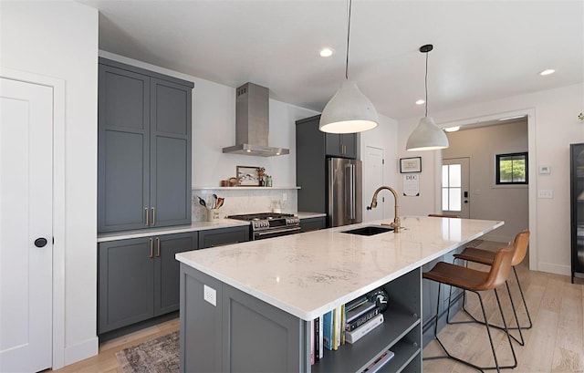 kitchen with hanging light fixtures, wall chimney range hood, light stone counters, a center island with sink, and appliances with stainless steel finishes