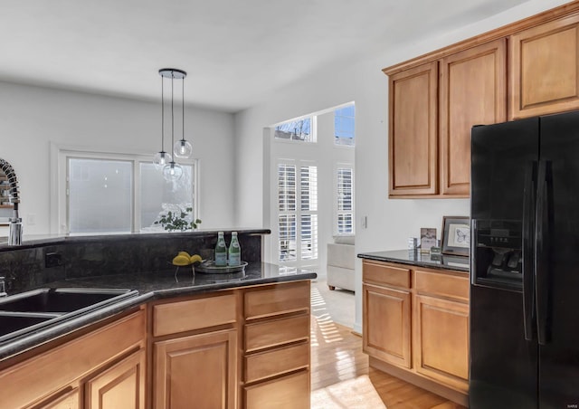 kitchen with light wood-type flooring, black fridge with ice dispenser, dark stone countertops, hanging light fixtures, and sink