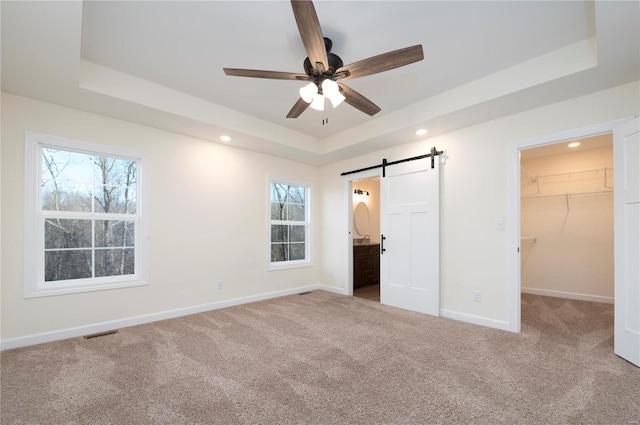 unfurnished bedroom featuring a raised ceiling, ensuite bath, ceiling fan, a barn door, and a spacious closet