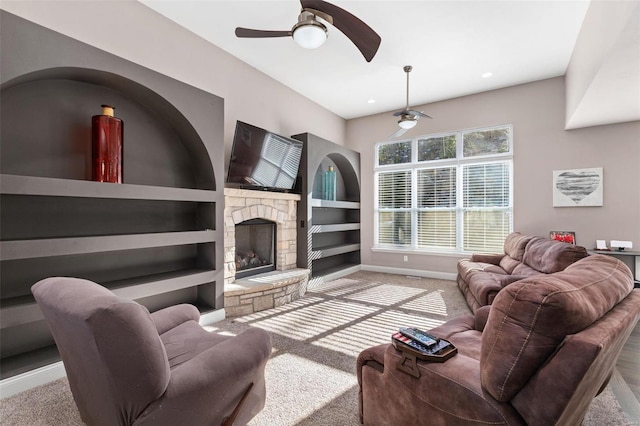 living room with a stone fireplace, light carpet, built in shelves, and ceiling fan