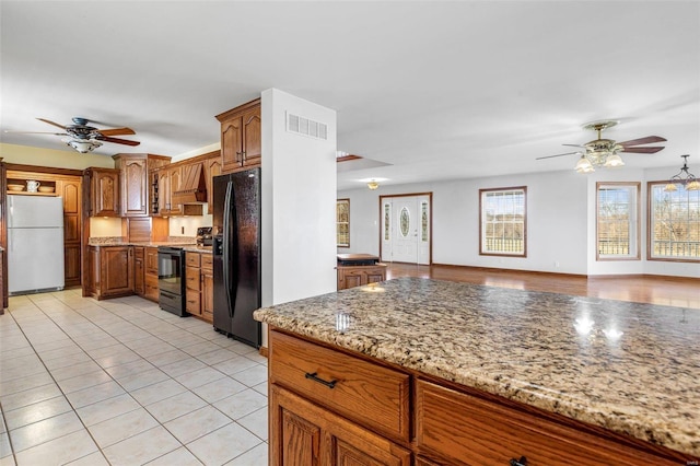 kitchen with light stone counters, light tile patterned flooring, black appliances, ceiling fan with notable chandelier, and custom exhaust hood