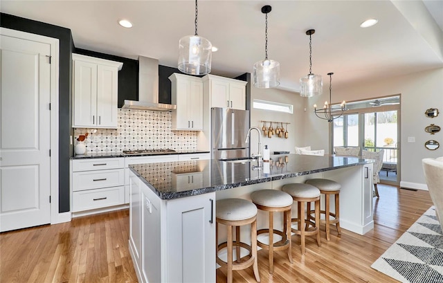 kitchen featuring wall chimney exhaust hood, an island with sink, pendant lighting, white cabinets, and appliances with stainless steel finishes