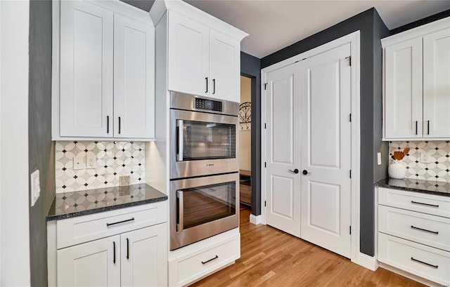 kitchen featuring white cabinetry, double oven, dark stone counters, decorative backsplash, and light wood-type flooring