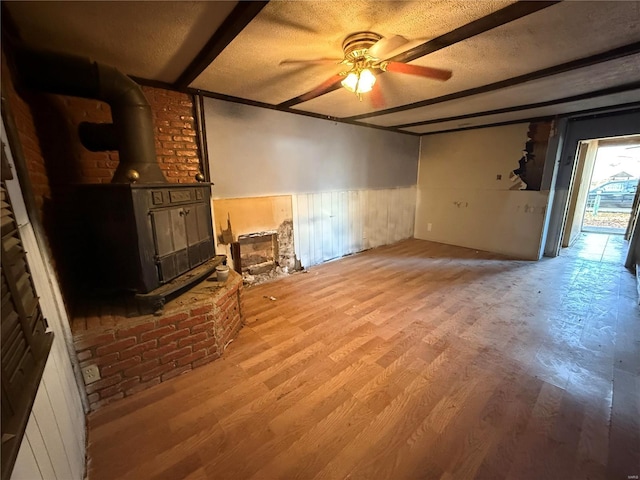 unfurnished living room with a textured ceiling, ceiling fan, wood-type flooring, beam ceiling, and a wood stove