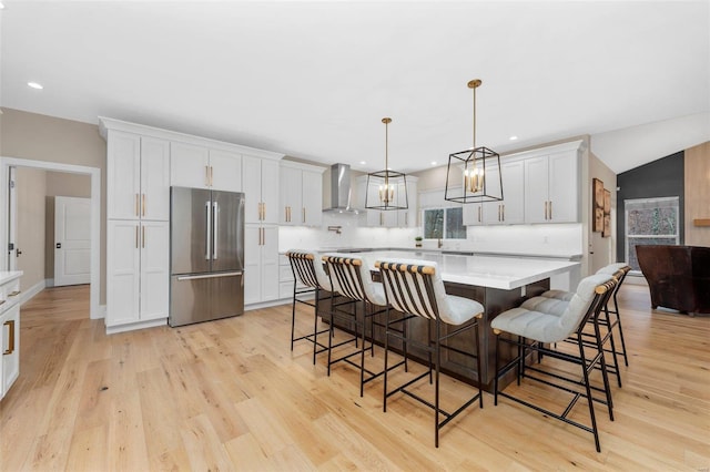 kitchen with stainless steel fridge, wall chimney exhaust hood, a spacious island, decorative light fixtures, and white cabinets