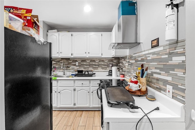 kitchen featuring sink, wall chimney exhaust hood, light hardwood / wood-style flooring, black refrigerator, and white cabinets