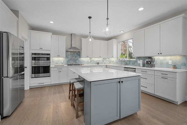 kitchen with white cabinetry, a kitchen island, wall chimney range hood, stainless steel appliances, and hanging light fixtures