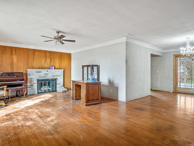 unfurnished living room with ceiling fan with notable chandelier, wooden walls, crown molding, light hardwood / wood-style flooring, and a stone fireplace