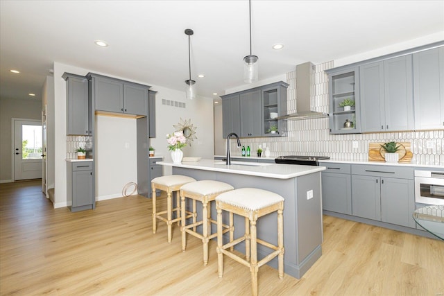 kitchen with gray cabinetry, a center island with sink, wall chimney range hood, sink, and decorative light fixtures